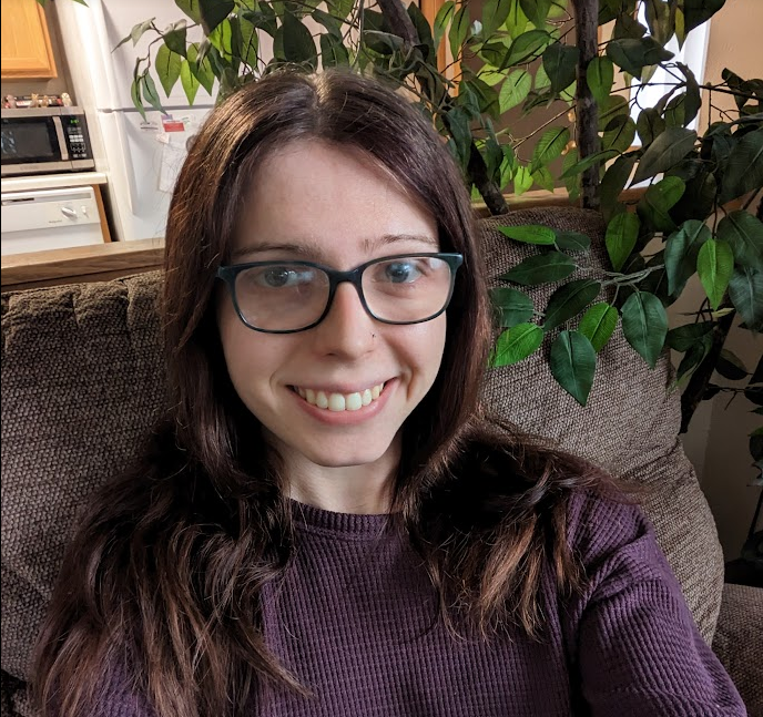 A white woman with long brown hair wearing glasses and a purple sweater smiling at the camera.